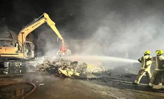 Firefighters spray water on a pile of smouldering rubbish, with plant machinery in the background