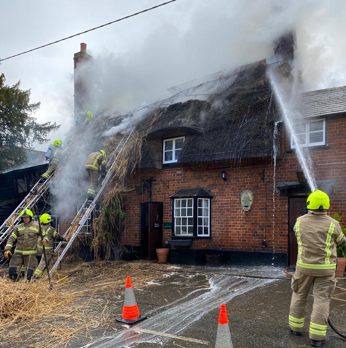 Six firefighters are tackling a fire at a house which is make of bricks and has a thatched roof. They are using ladders and hose reel jets. There is smoke coming from the roof.