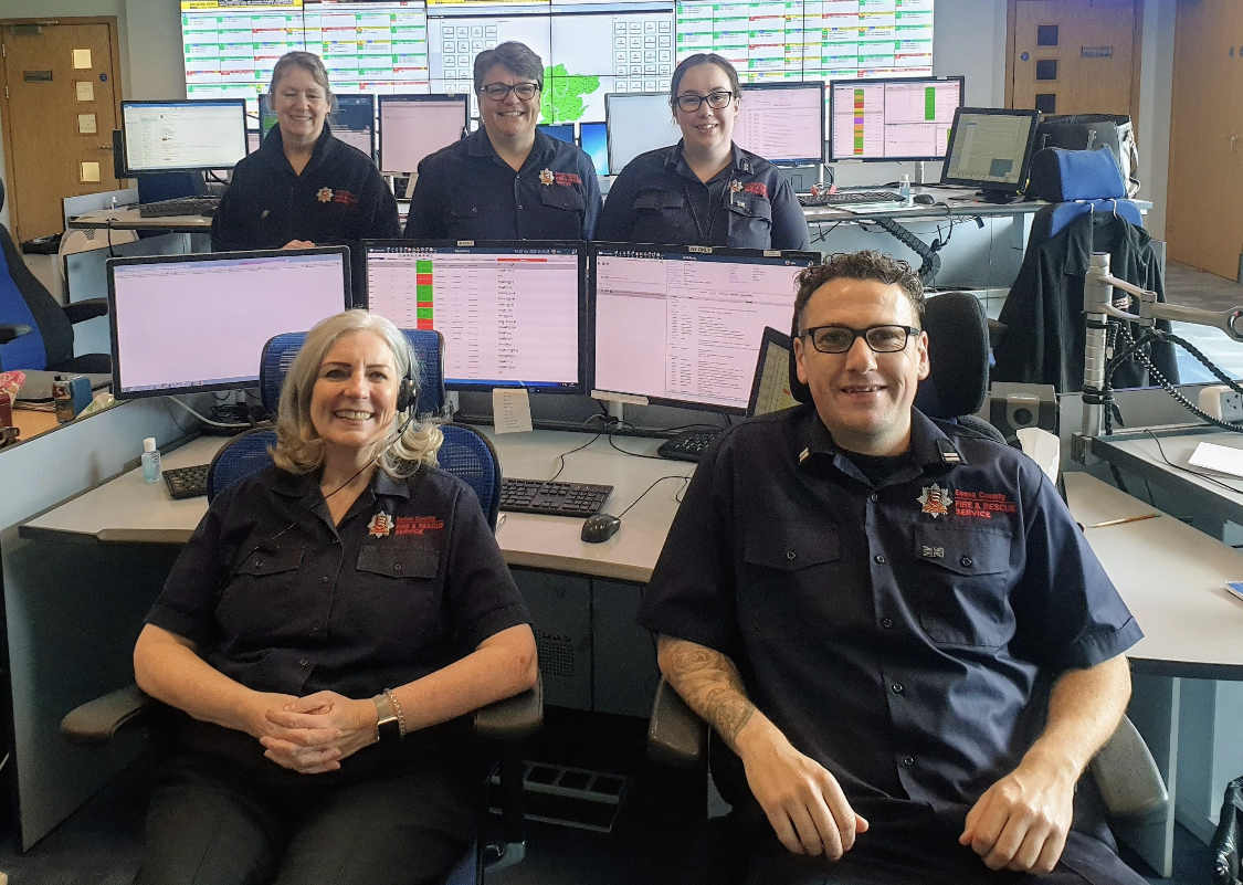 Five control room operators all in blue uniform. Two are sat at a desk, a man and woman, and three women are stood behind some screens behind them. They are all smiling.