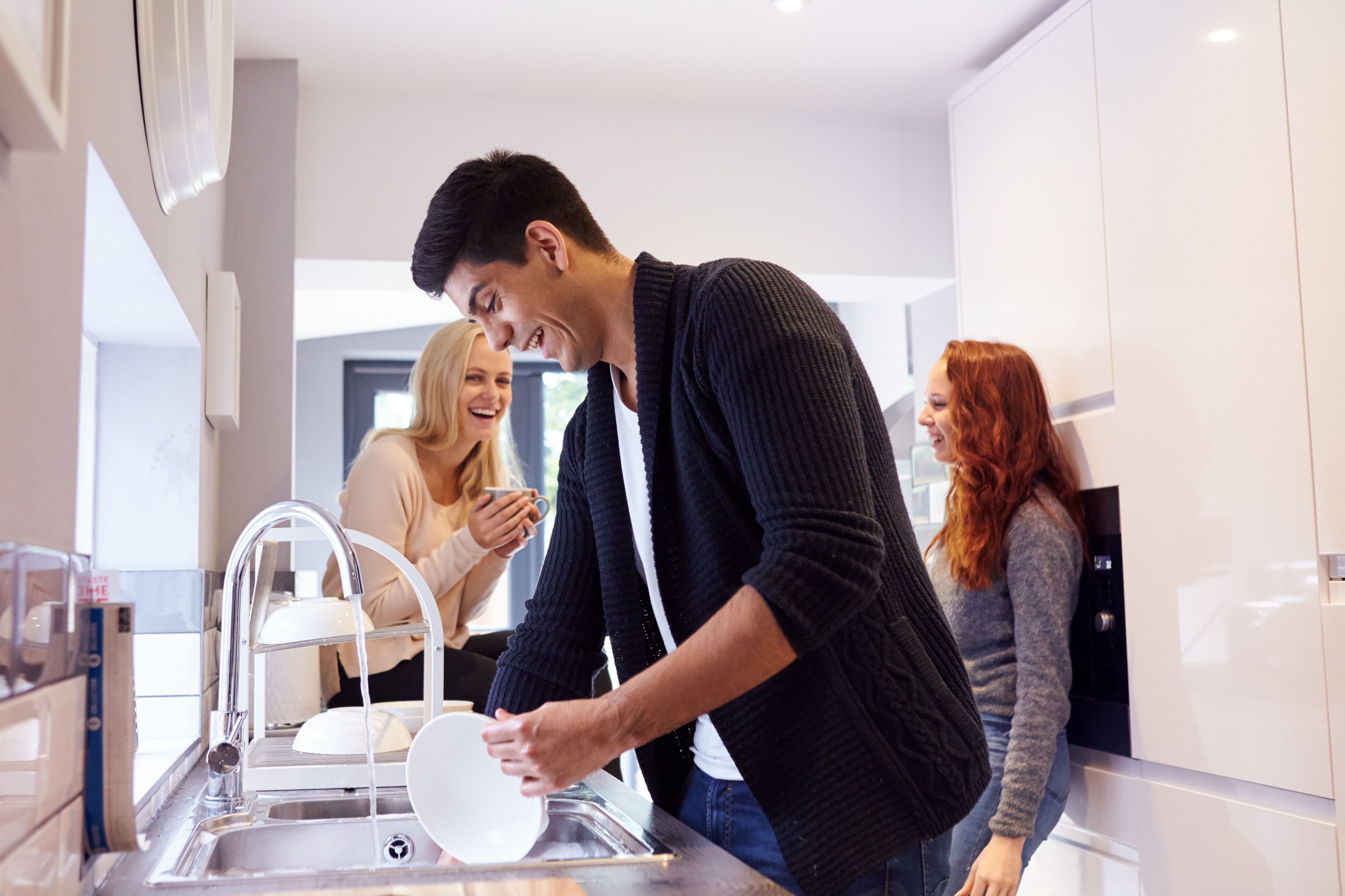 Three students in the kitchen. A young man is filling a jug with water and two girls are smiling at each other in the background