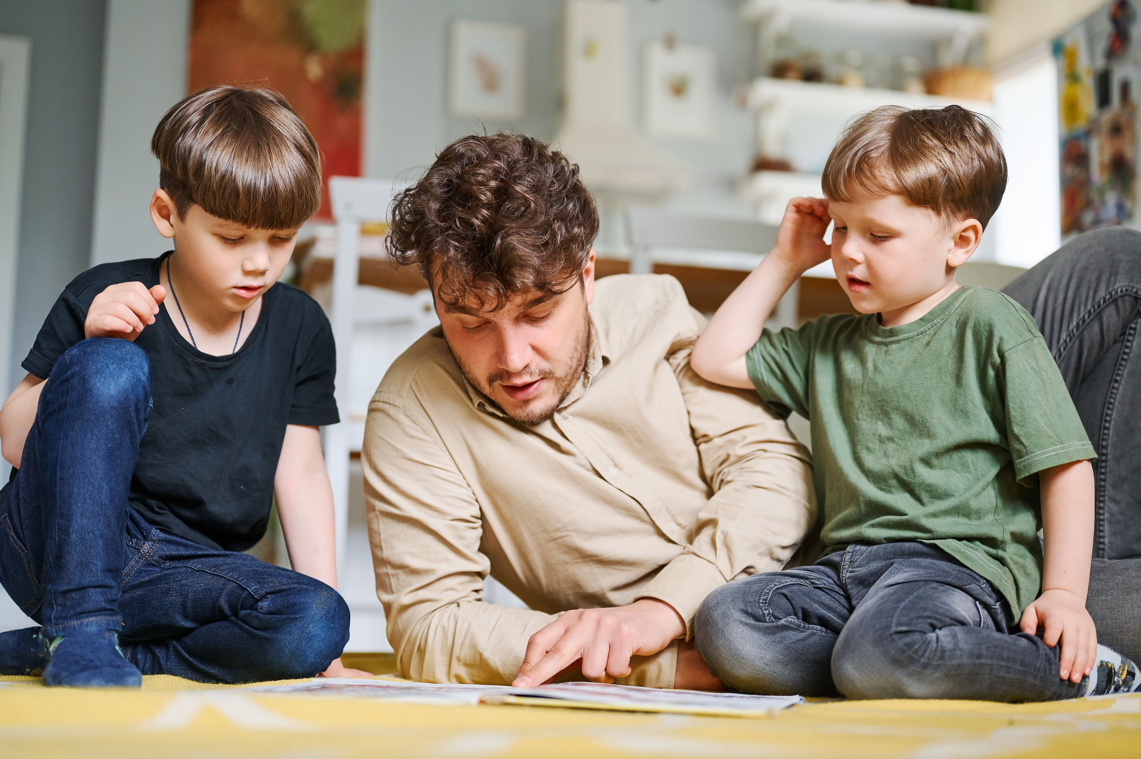 Dad and two young boys sat on a wooden floor, looking at a book together