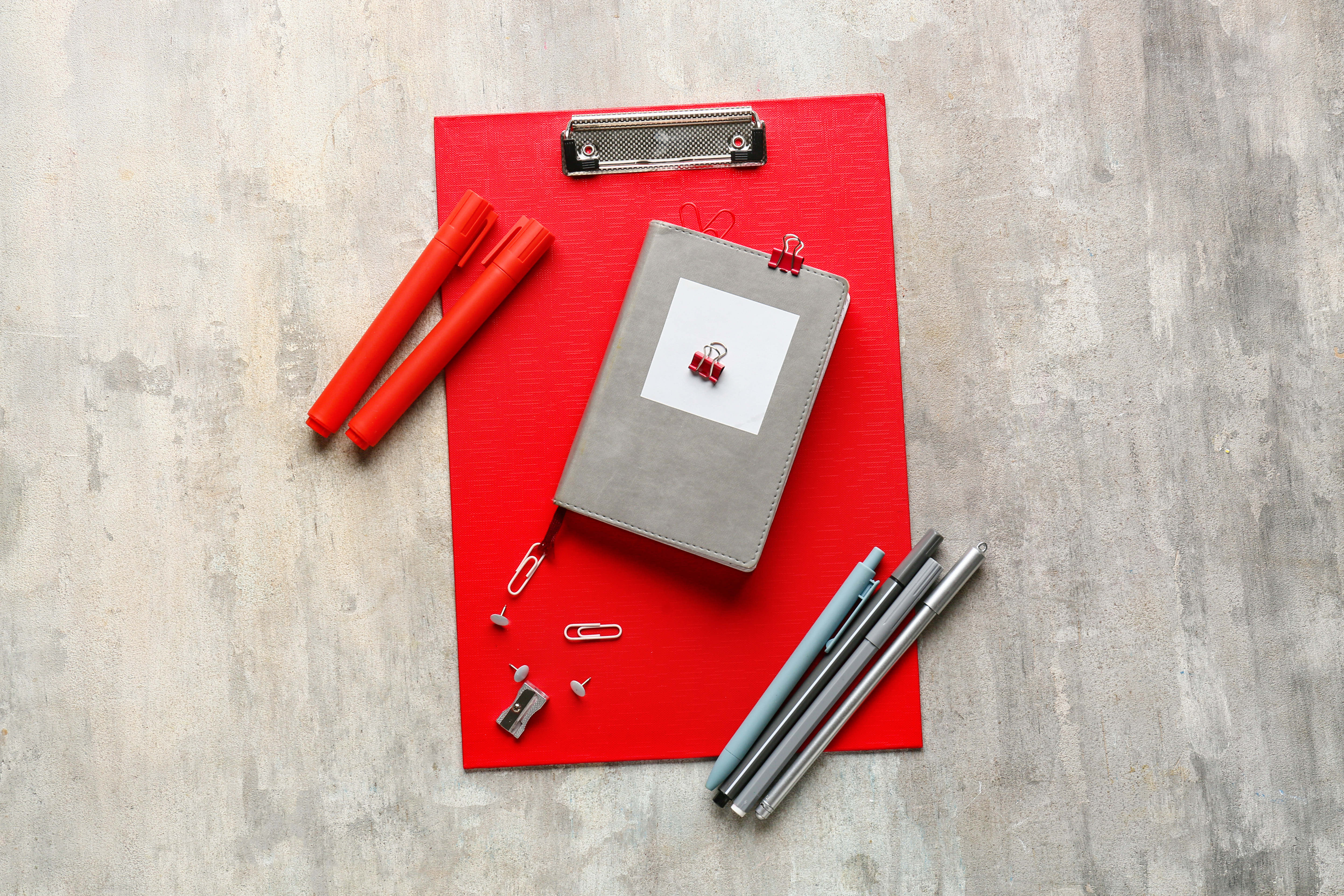 Red clipboard, notepad and pens spread across a textured grey table