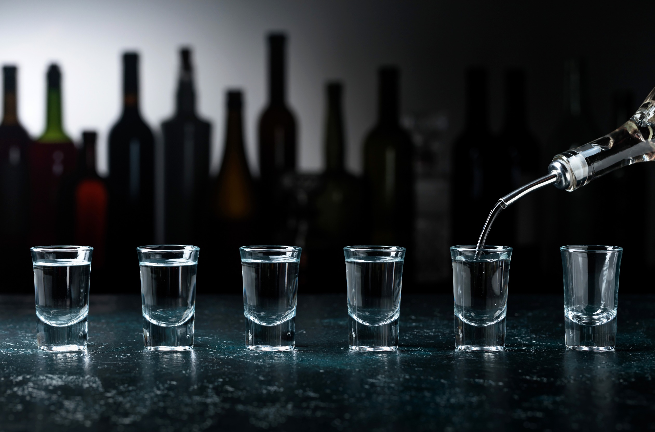 Bottle of spirit being poured into six shot glasses on bar