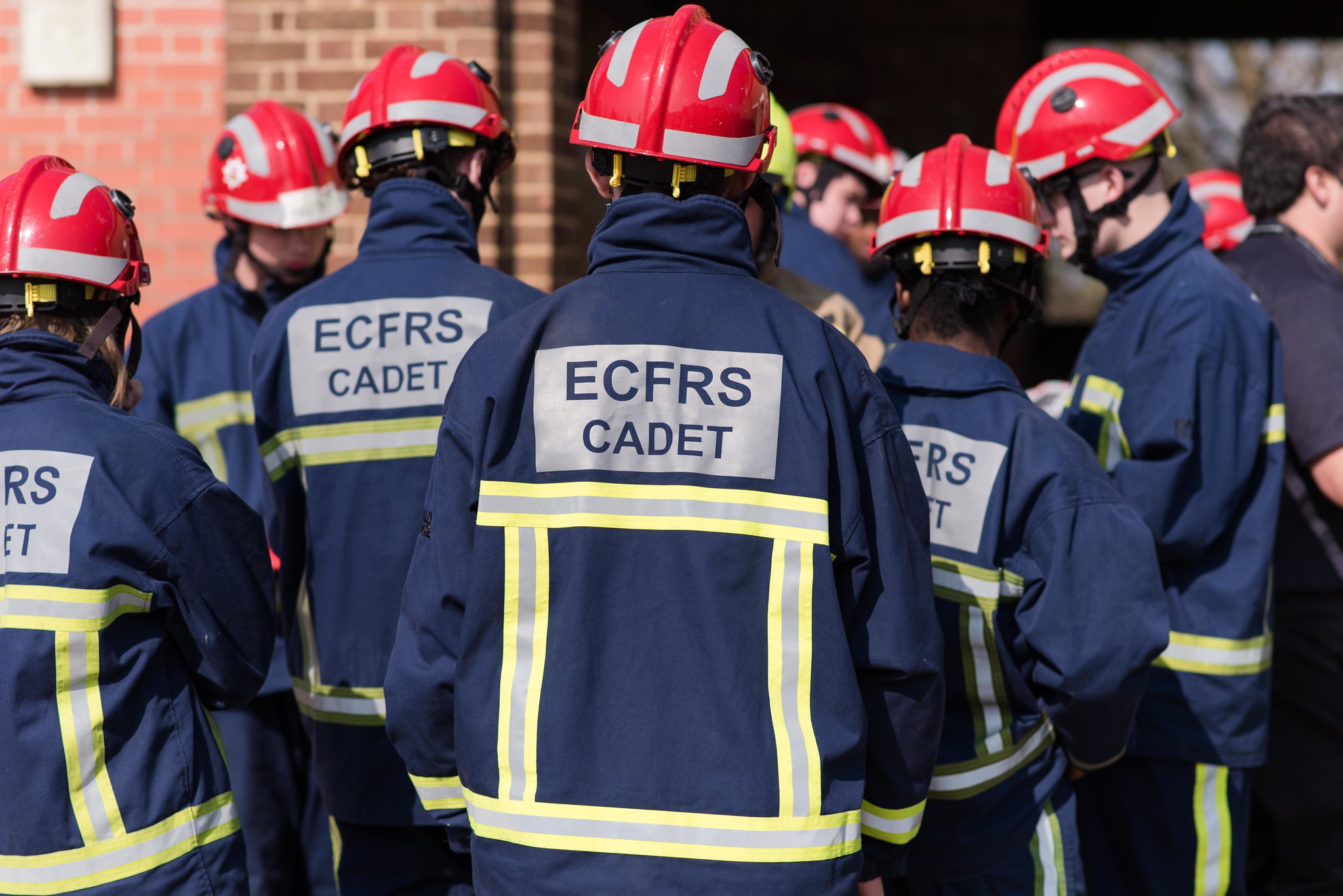 A group of teenage fire cadets pictured from behind, wearing navy PPE and red helmets