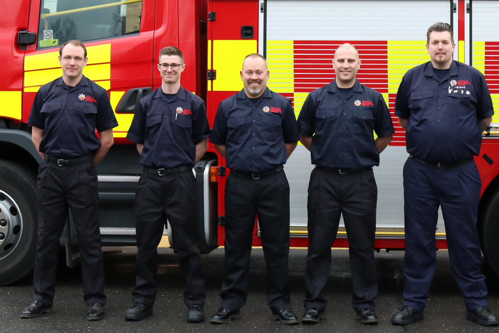 Five firefighters standing in front of a fire engine and smiling into the camera.