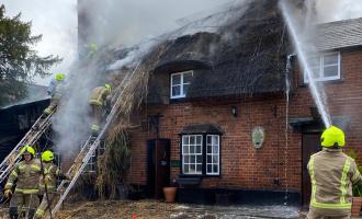 Six firefighters are tackling a fire at a house which is make of bricks and has a thatched roof. They are using ladders and hose reel jets. There is smoke coming from the roof.