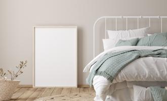 Beige bedroom with natural floor. White bed frame with multiple white and blue blankets layered on top. A mirror is propped up against the wall.