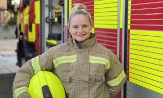 Photo of a female firefighter standing in front of a fire appliance. She is holding her helmet under her arm. 