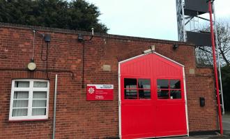 Small brick fire station building with one red shutter door. Drill tower visible is visible in the background.