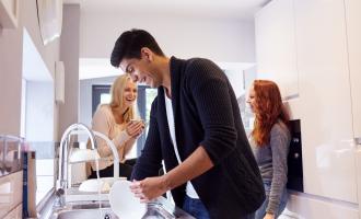 Three students in the kitchen. A young man is filling a jug with water and two girls are smiling at each other in the background