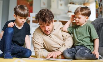 Dad and two young boys sat on a wooden floor, looking at a book together