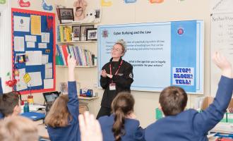 Education Officer standing by a projector in front of primary school children in blue uniform. One boy has raised his hand