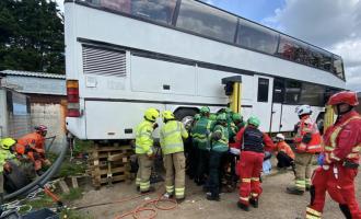 A white double decker bus lifted with a large group of people around it, attempting to release a man who is trapped near the wheel. 