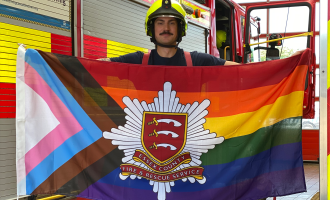 Firefighter Matt Hill holding the progress rainbow flag with the ECFRS crest in the centre. Matt is waering a firefighter helmet and standing in front of a fire engine. 