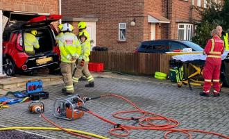 Firefighters working next to and inside a car, which has gone through the front of a house, as paramedics look on