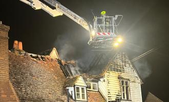 A firefighter standing on an aerial ladder platform in front of a fire damaged roof