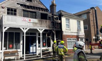 Wooden building (sailing club) damaged by fire with firefighters in shot.