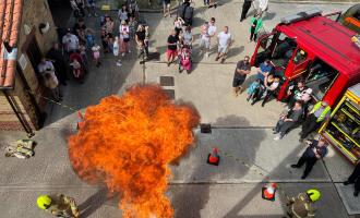 A drill yard at a fire station, full of people for an event, a fire engine and a large fire from a demonstration