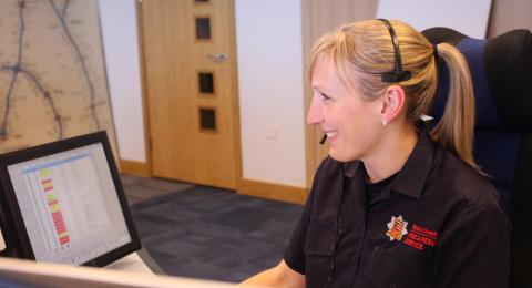 A female control room operator taking a call in our control room. She is smiling and sitting in front of a computer screen and is wearing a headset.