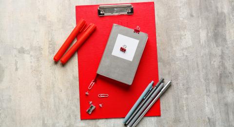 Red clipboard, notepad and pens spread across a textured grey table