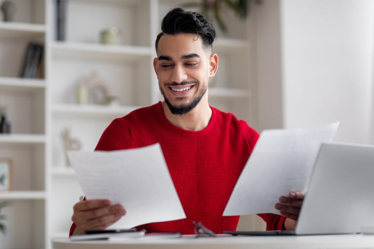 Man in red jumper holding two pieces of paper sat at white table with laptop