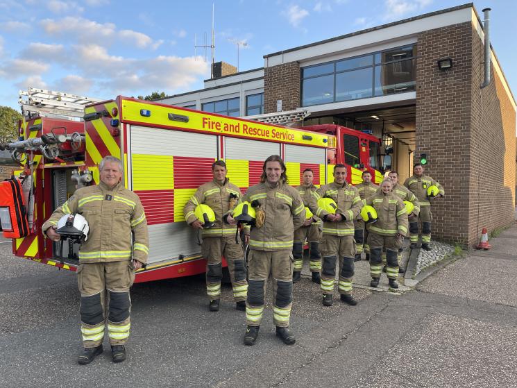 This is an image of eight firefighters, men and women, standing in their protective clothing in front of a fire engine. They are all smiling and holding their helmets in their arms.