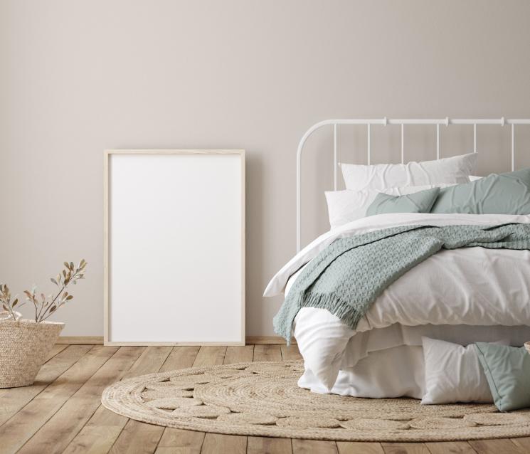 Beige bedroom with natural floor. White bed frame with multiple white and blue blankets layered on top. A mirror is propped up against the wall.