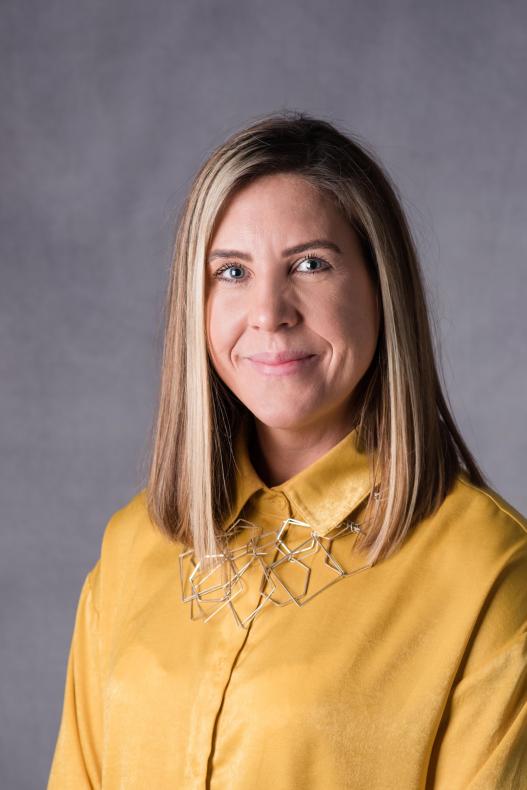 Head and shoulders photo of Emily Cheyne, Assistant Director, Communications, Marketing and Brand. Emily is smiling and wearing a mustard yellow shirt. She has shoulder length hair.