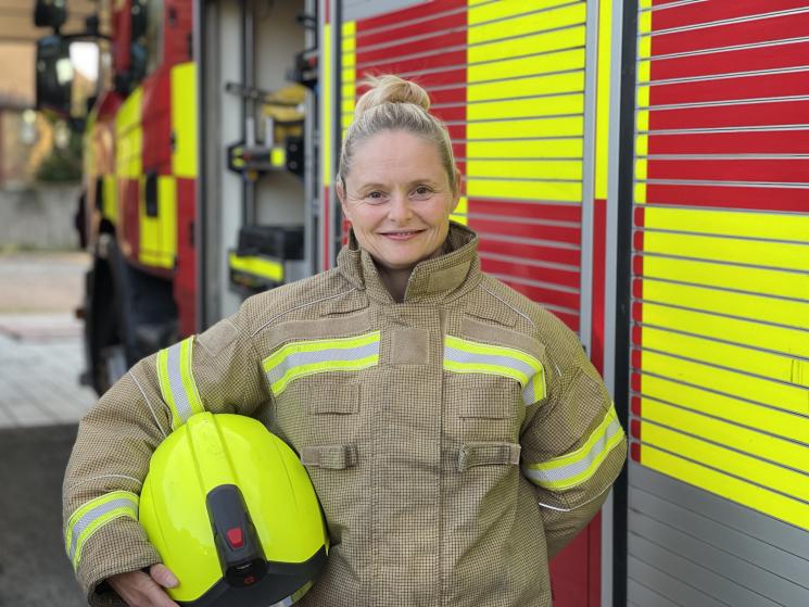 Photo of a female firefighter standing in front of a fire appliance. She is holding her helmet under her arm. 