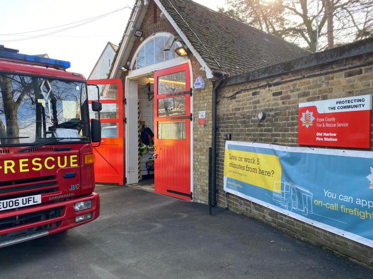 Small brick fire station building with one red shutter door. Fire Engine parked outside the front of building.