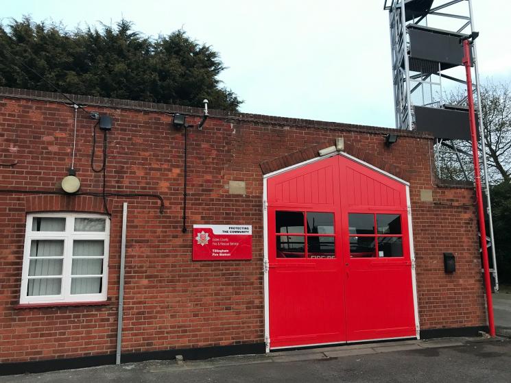 Small brick fire station building with one red shutter door. Drill tower visible is visible in the background.