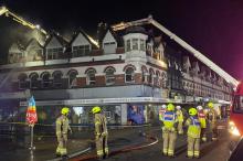 Firefighters standing outside a three storey building, with two Aerial Platform Ladders tackling a fire