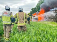 Firefighters spraying water on the woodpile and vehicles, with two fire officers in the foreground