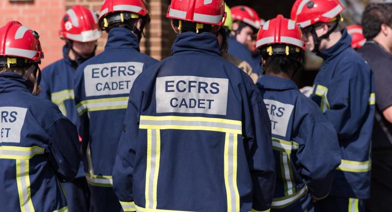 A group of teenage fire cadets pictured from behind, wearing navy PPE and red helmets