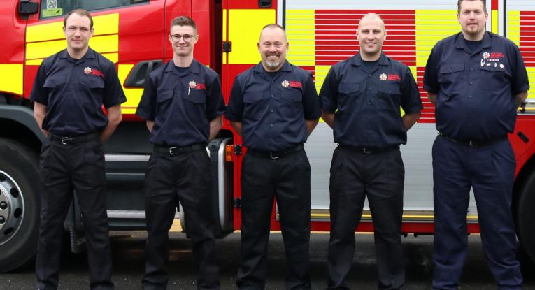 Five firefighters standing in front of a fire engine and smiling into the camera. 