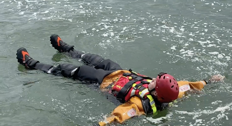 A firefighter floating on his back with his arms and legs stretched out in water to demonstrate how to float to live